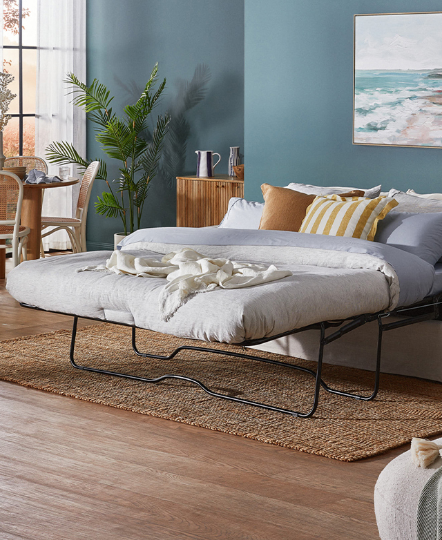 The sofa-bed is unfolded into a queen-sized bed, which is dressed in neutral bed sheets and golden cushions. The sofa-bed is unfolded into a queen-sized bed, which is dressed in neutral bed sheets and golden cushions.