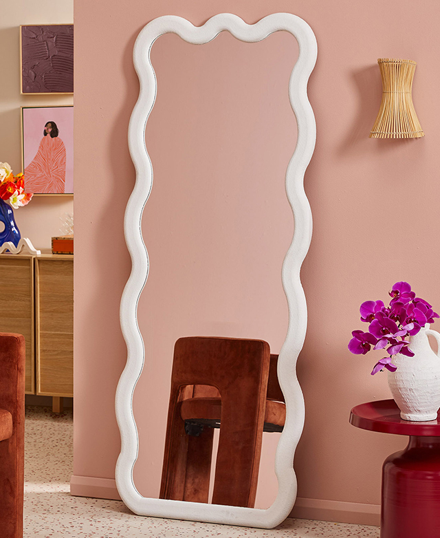 The tall, white wavy mirror is leaning against a pink wall in a bright room that features terrazzo floors and art. The tall, white wavy mirror is leaning against a pink wall in a bright room that features terrazzo floors and art.