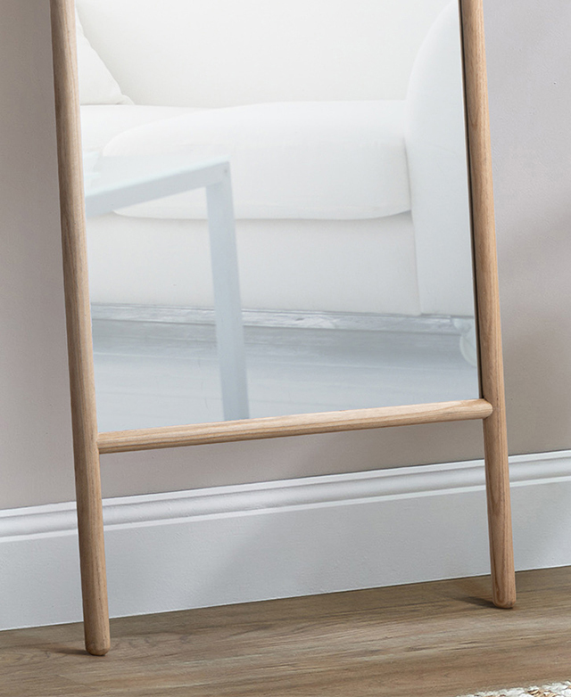Mirror is leaning against a soft pink wall with white skirting board; the glass reflects a sofa.