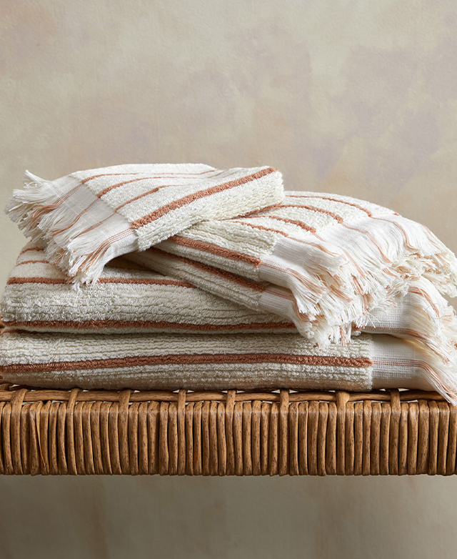 A stack of clay and cream striped bath towels with fringing is on a jute stool.