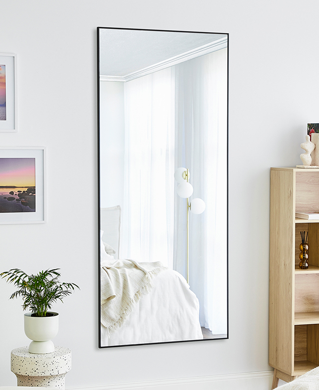 The rectangular black-framed mirror is affixed to a white wall in a bright room, where artwork and a plant is near. The rectangular black-framed mirror is affixed to a white wall in a bright room, where artwork and a plant is near.