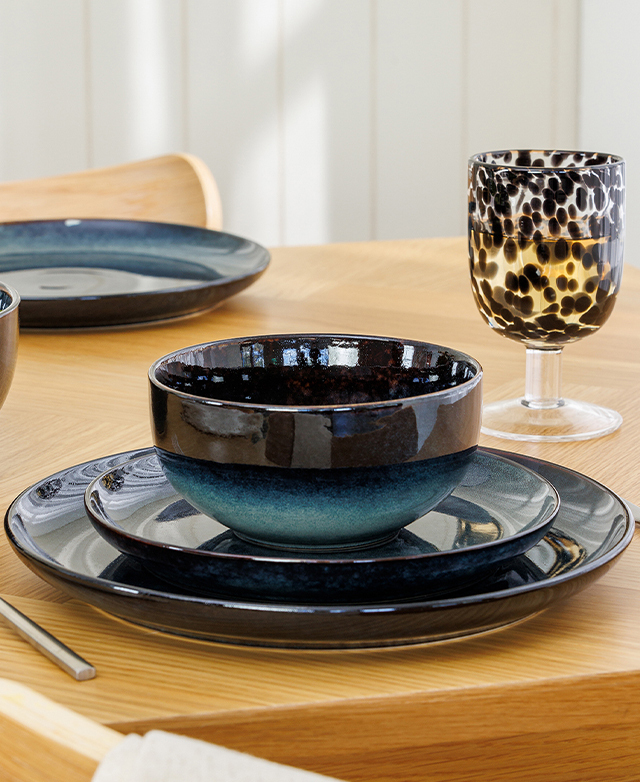 A timber table is topped with layered blue crockery, with a brown Murano-style glass next to it.