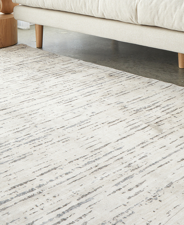The cream, blue, and charcoal abstract rug is positioned on a concrete floor in front of a cream sofa. The cream, blue, and charcoal abstract rug is positioned on a concrete floor in front of a cream sofa.