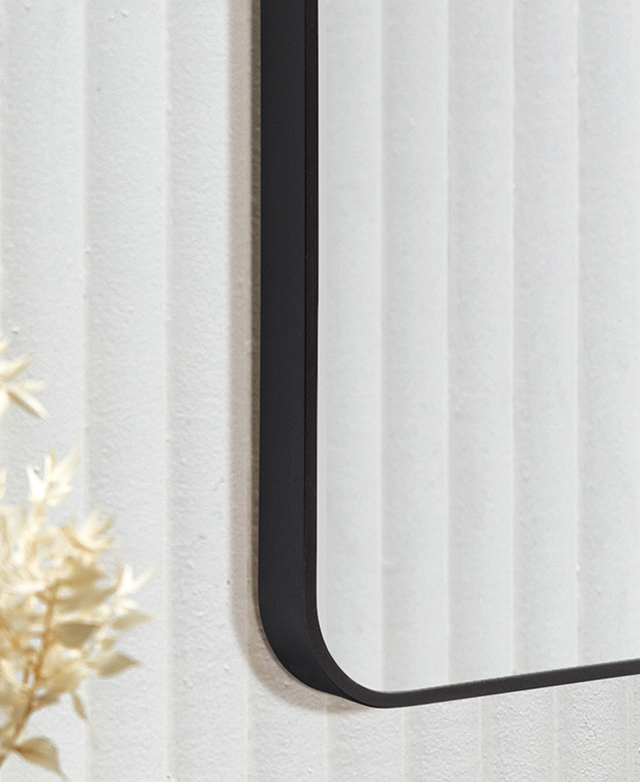 The mirror's slender black metal frame has curved corners, and one is shown against a white fluted wall.