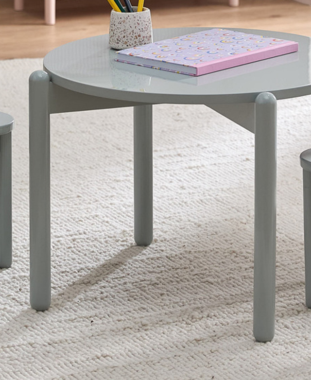 A rainbow book and ceramic pencil holder is on top of the round, glossy blue children's table, which is on a cream rug.