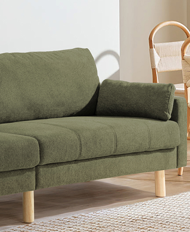 The green sofa is shown in a bright living room, with a jute rug in front of it, and rattan dining chairs in the background.