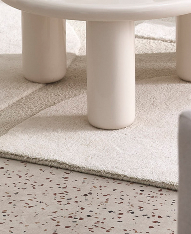 The cutwork cream rug is laid on a speckled concrete floor, with a glossy white coffee table on top.