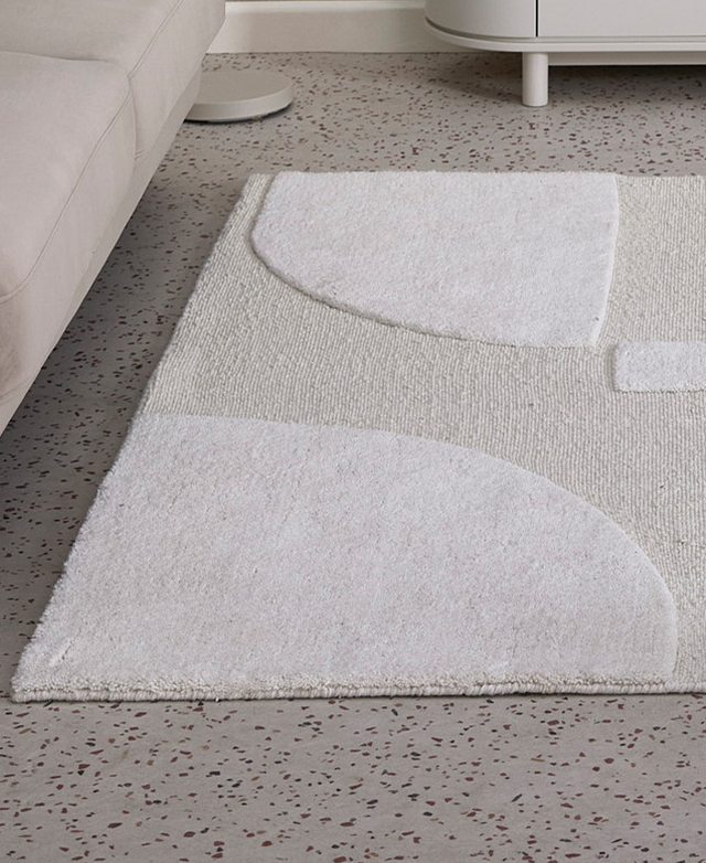 The textured, geomteric cream rug is pictured on a speckled concrete floor in front of a cream rug.