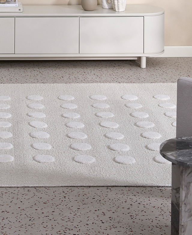 The textured, spotted cream rug is depicted atop a speckled concrete floor in a bright room next to a console table. The textured, spotted cream rug is depicted atop a speckled concrete floor in a bright room next to a console table.