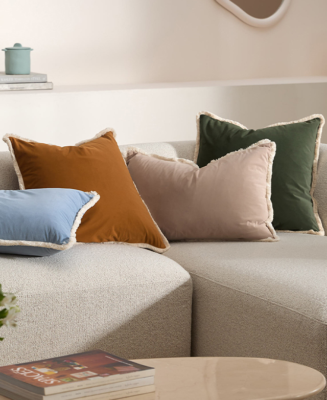 Four differently coloured velvet cushions are positioned atop a cream sofa, which has a coffee table in front of it.