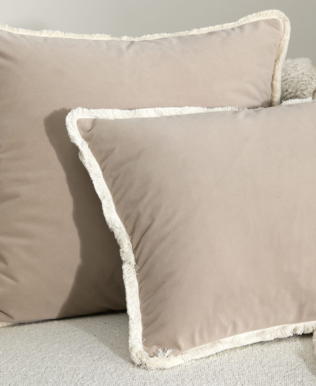 A close-up of two taupe velvet cushions with fringed trims are on top of a cream sofa.