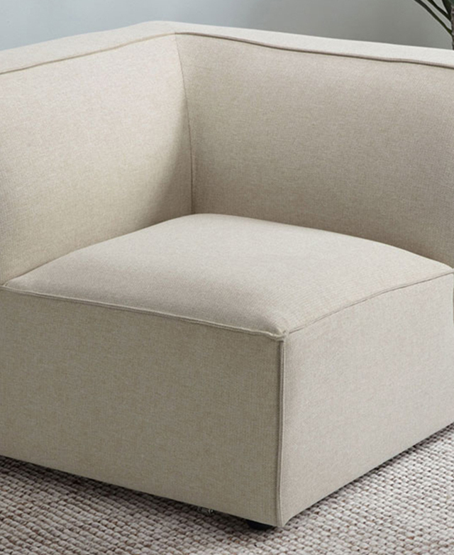 A cream corner chair has clean lines, a thick backrest, and piping trims; it's on a cream woven rug. A cream corner chair has clean lines, a thick backrest, and piping trims; it's on a cream woven rug.
