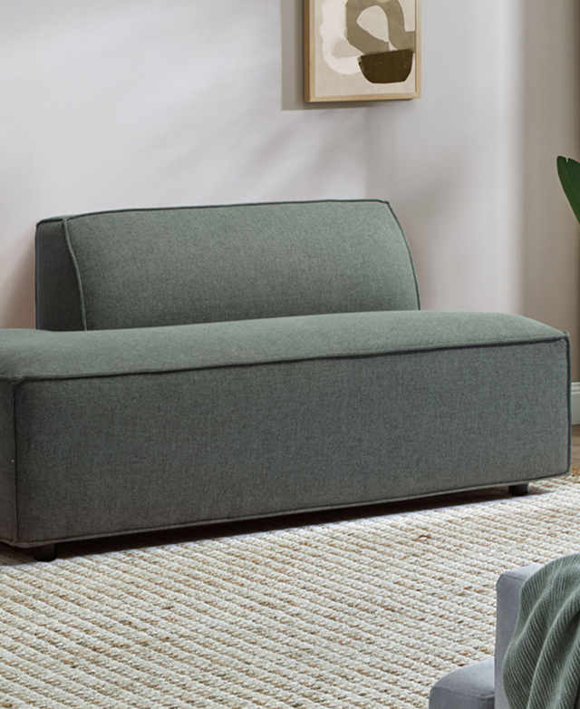 The chaise section of the green modular sofa is flush against a soft grey wall, on top of a natural jute rug.