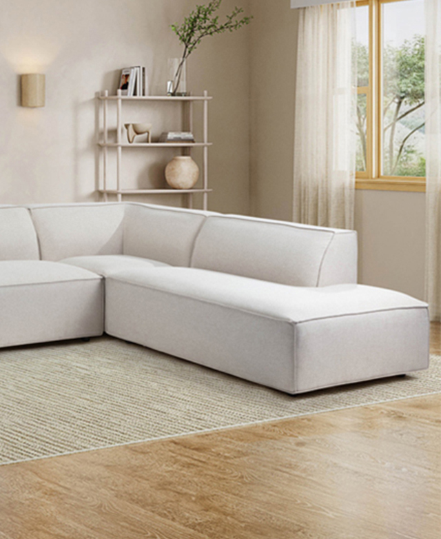 The cream chunky L-shaped sofa is in a warm, bright room on top of a taupe rug. A bookshelf is in the background.