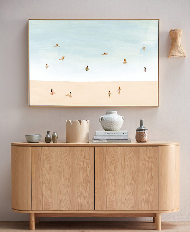 The beach scene wall art hangs over a modern pill-shaped console, which is topped with various decorative items.