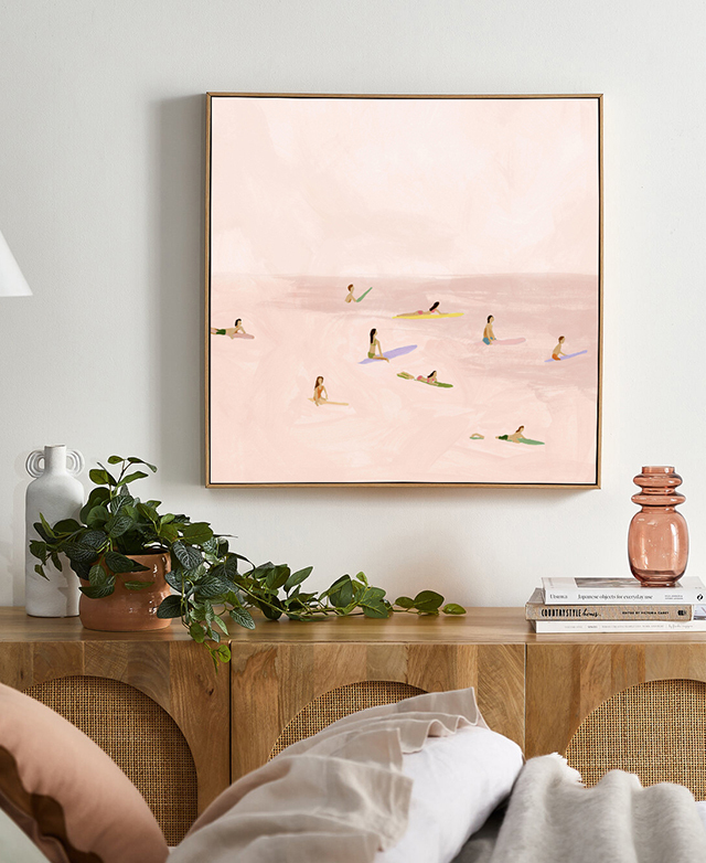 The pink-toned wall art hangs over a console table that has rattan panelled cupboards, and is topped with a plant and decor. The pink-toned wall art hangs over a console table that has rattan panelled cupboards, and is topped with a plant and decor.