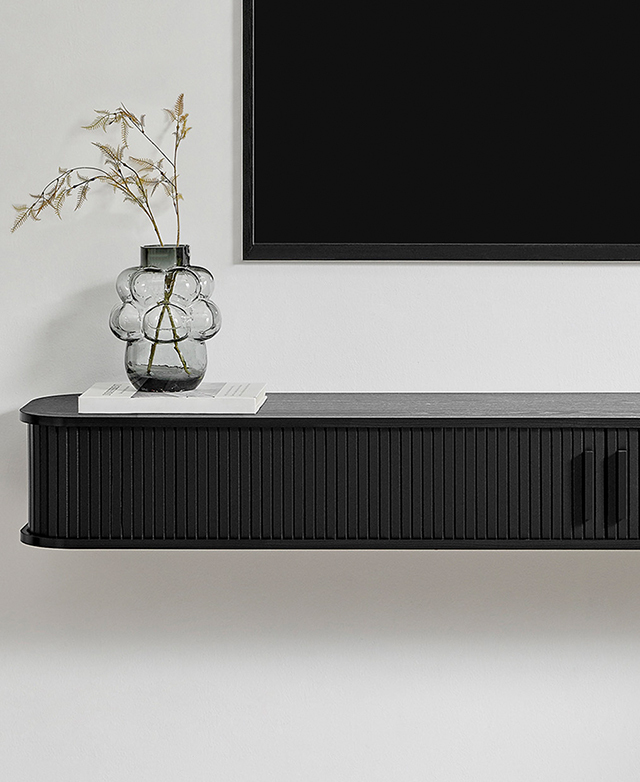 The black floating TV unit is affixed to a white wall and styled with a stack of books with a large glass vase on top.