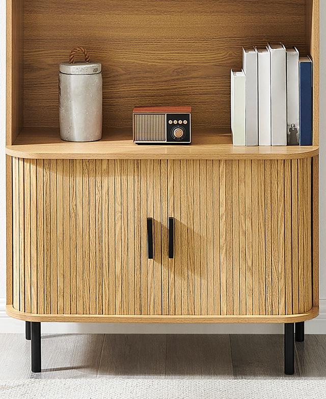 Books, decor, and a radio adorn the bottom shelf of the fluted, oak cabinet. Books, decor, and a radio adorn the bottom shelf of the fluted, oak cabinet.