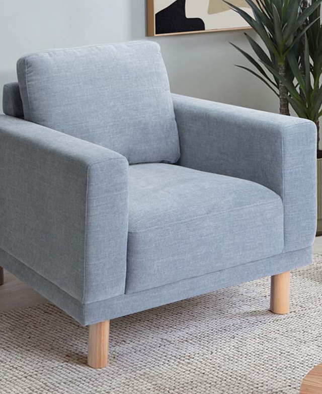 The blue armchair is in a soft blue room, depicted next to a leafy plant and on top of a cream rug.