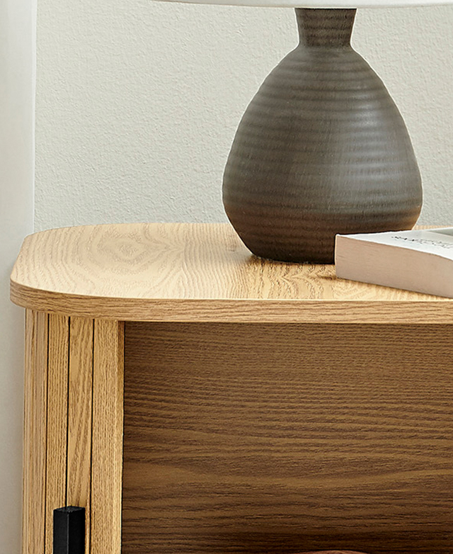 Cropped view of the side table shows the warm laminated surfaces on the inside and outside. A lamp and book is on top. Cropped view of the side table shows the warm laminated surfaces on the inside and outside. A lamp and book is on top.