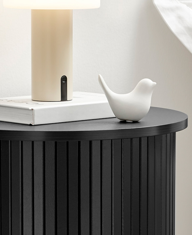 A lamp, book, and ceramic bird are on top of the black side table, which has smooth, laminated surfaces. A lamp, book, and ceramic bird are on top of the black side table, which has smooth, laminated surfaces.