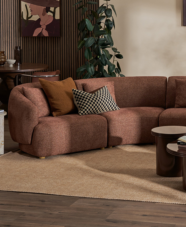 The rust-coloured modular sofa is shown on top of a caramel rug, with textured cushions, in a warm wood-panelled room.